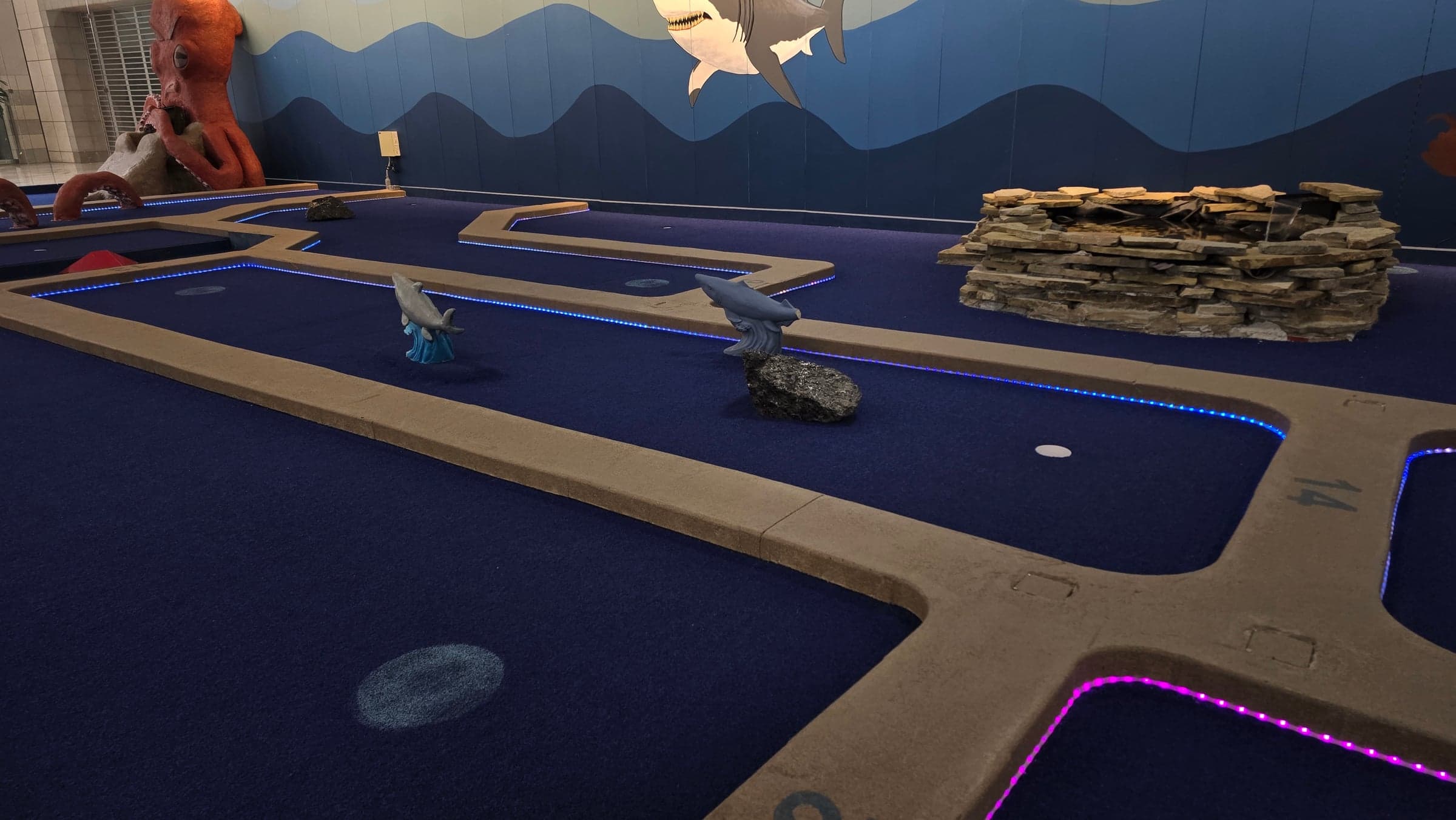 Blue and pink LED borders run around mini-golf holes with shark sculptures and a stone water feature.