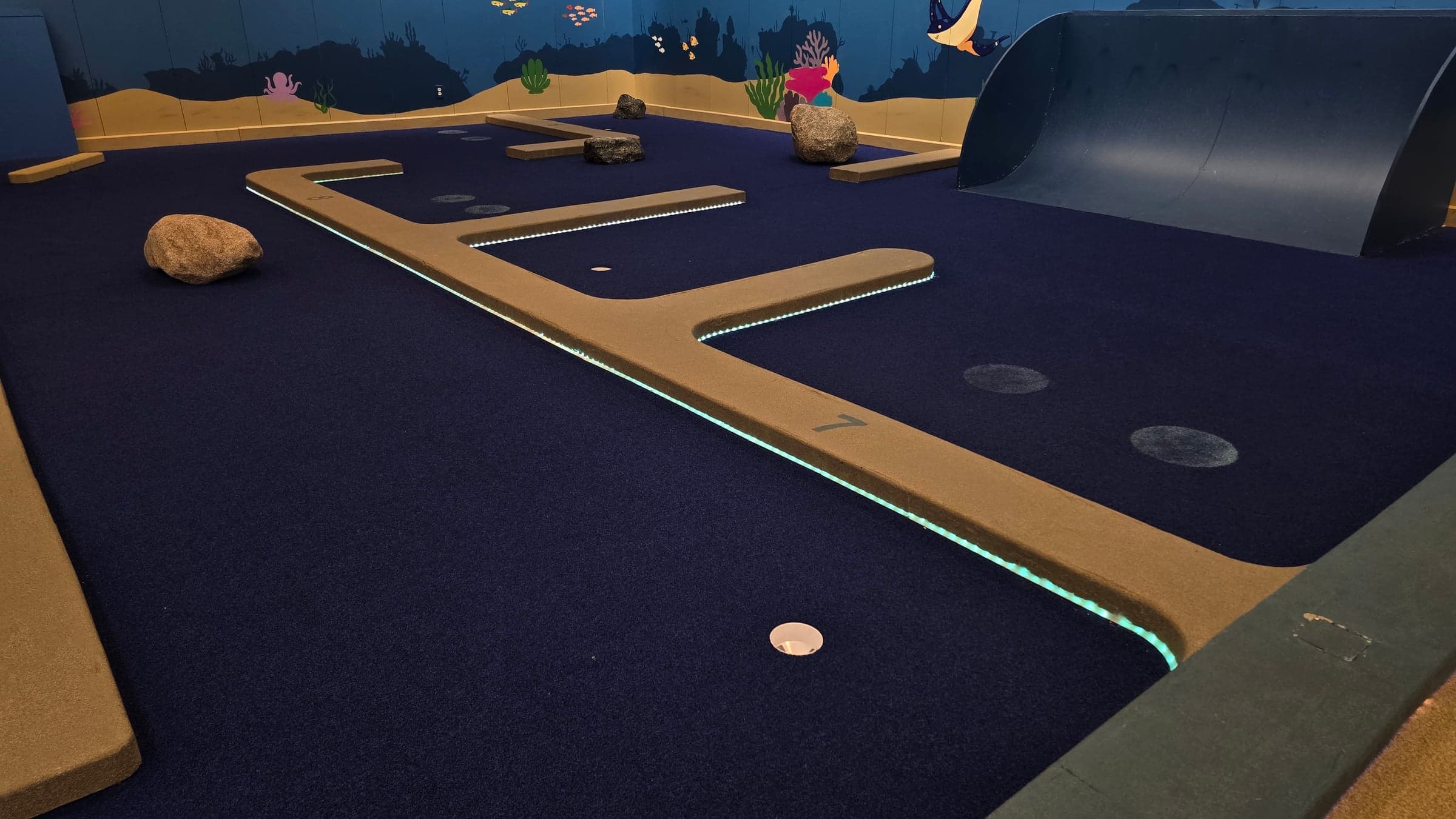 Teal LED strips illuminate the long border of a mini-golf hole with ocean murals and a curved ramp.