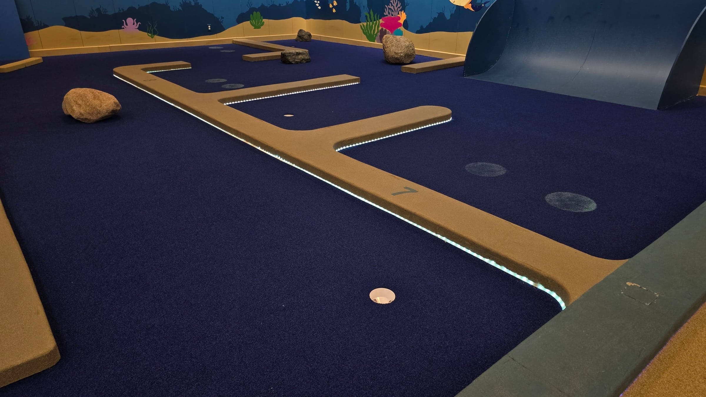 White LED strips follow the angular borders of a mini-golf hole beside a curved ramp.